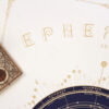 Close-up of the gold Ephemeris 2027 lettering on textured cream paper, beside antique astronomical instruments.