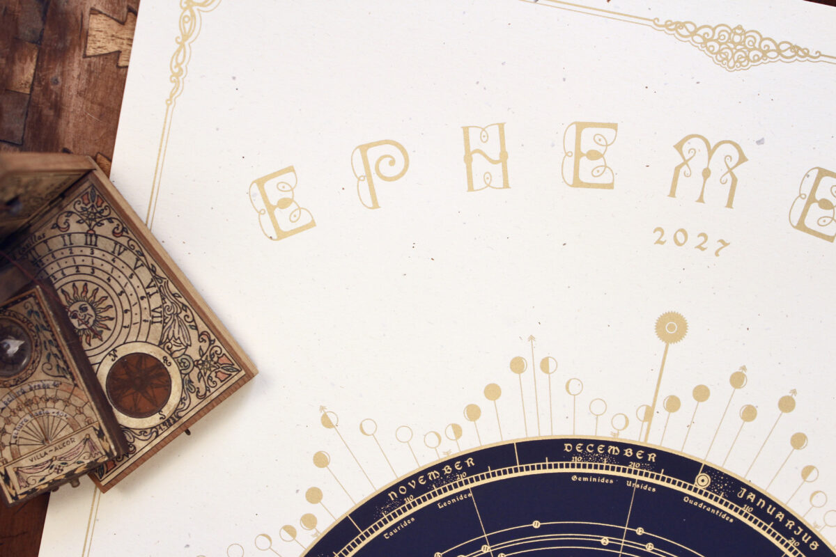 Close-up of the gold Ephemeris 2027 lettering on textured cream paper, beside antique astronomical instruments.