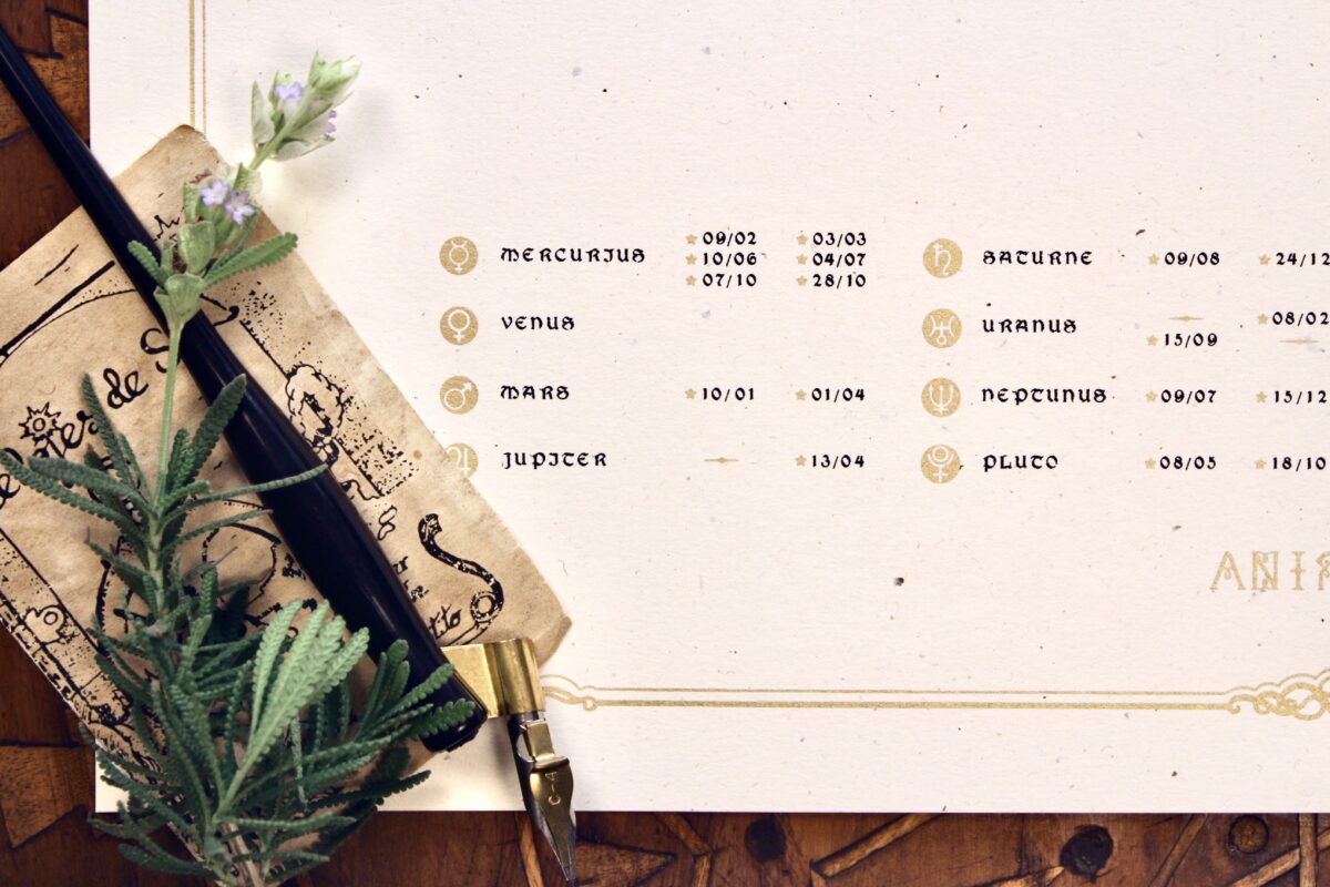 Close-up of planetary symbols and dates printed in gold on textured cream paper, beside a vintage-style pen and flowers.
